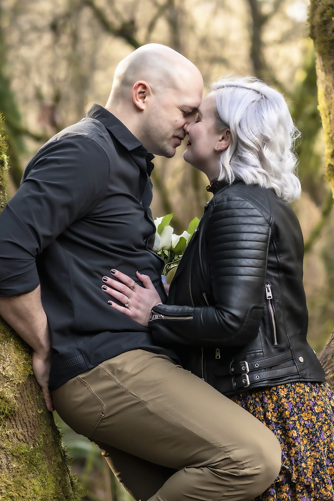 Man leans against a tree while a woman wearing a black leather jacket leans in. Both have their eyes closed and look ready to kiss. 