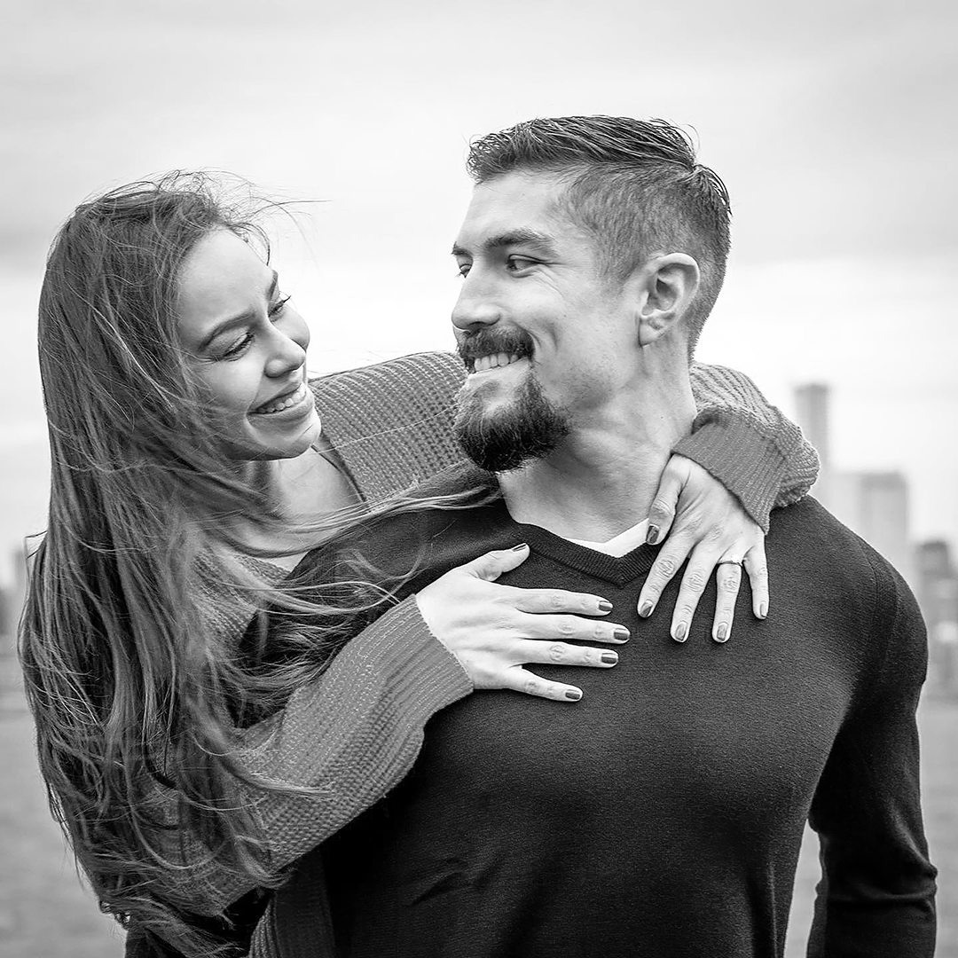 A couple looks into each others eyes with a city landscape behind them.