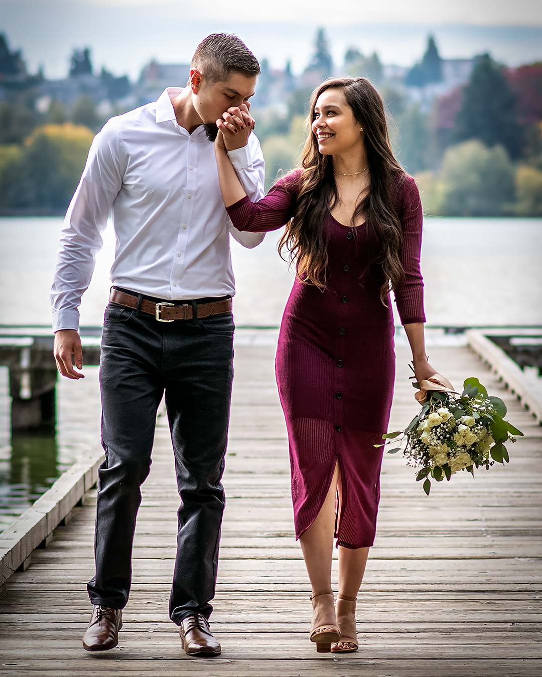 Man kisses his fiancé's hand while they walk down a wooden dock. 