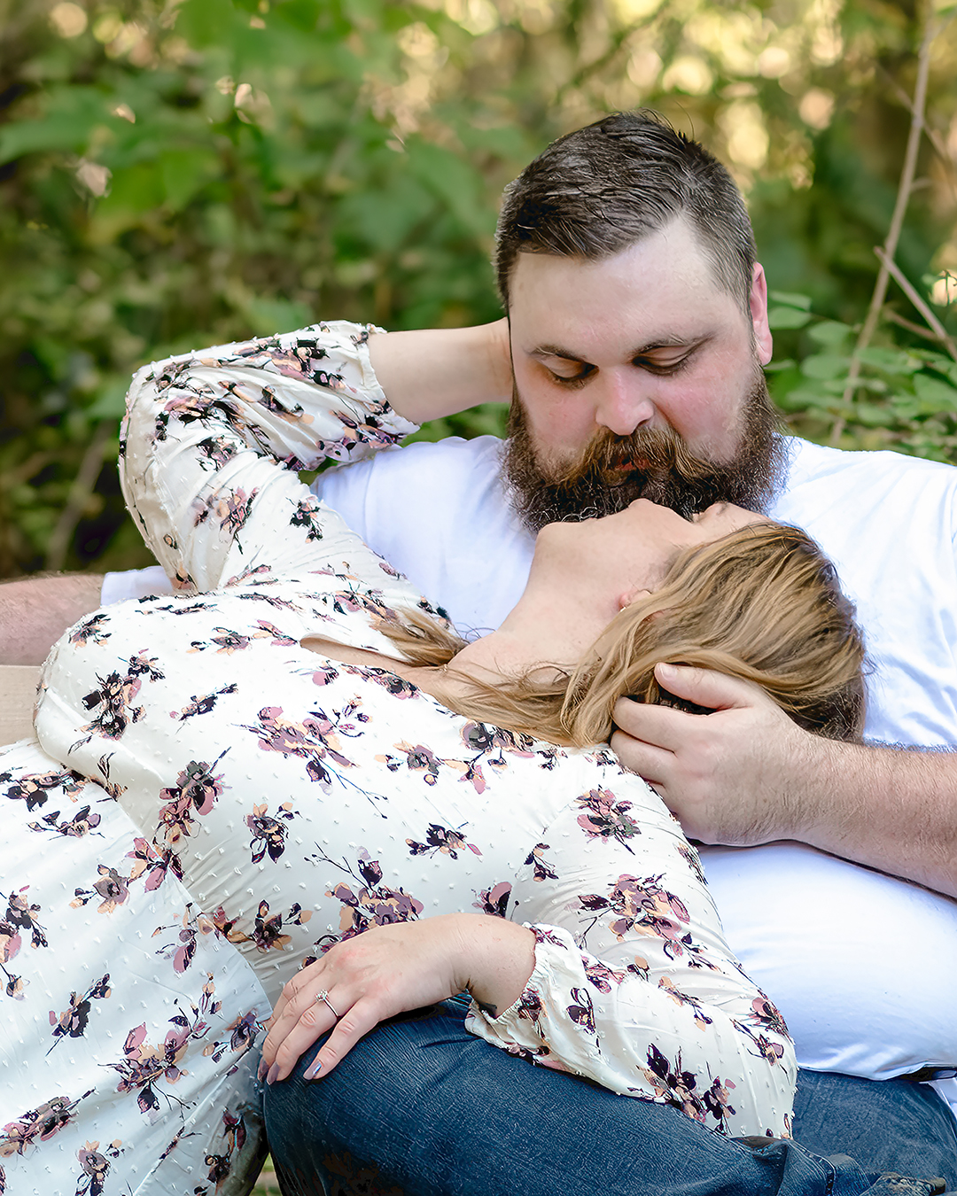 Woman lies in her fiancé's lap while sitting on a bench in the woods. 