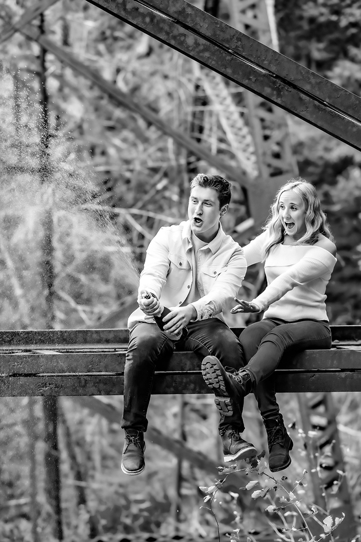 Black and white photo of a couple celebrating their engagement on a trestle bridge. 