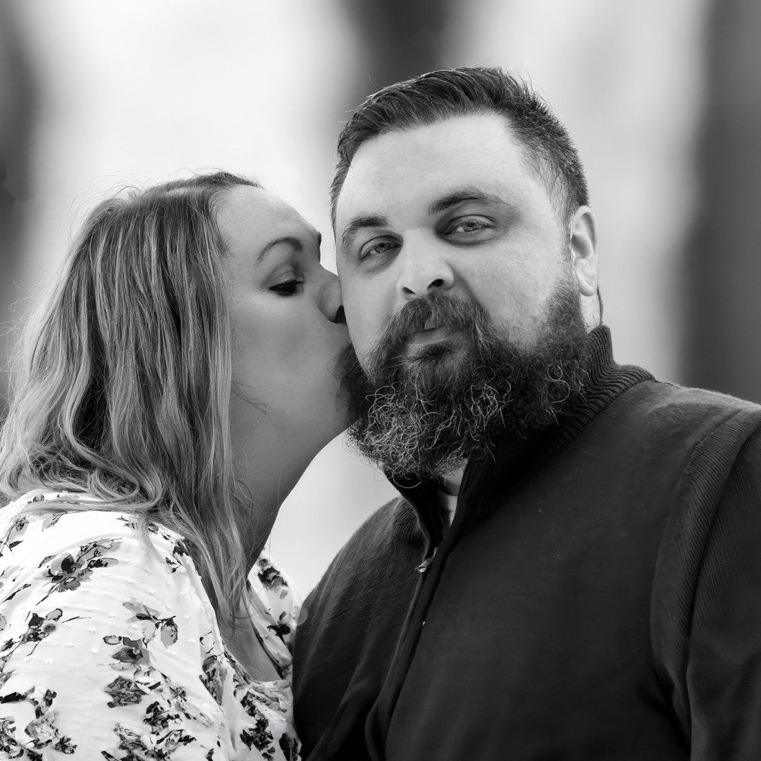 Kissing the cheek of her fiancé with a waterfall in the background. The photo is black and white. 