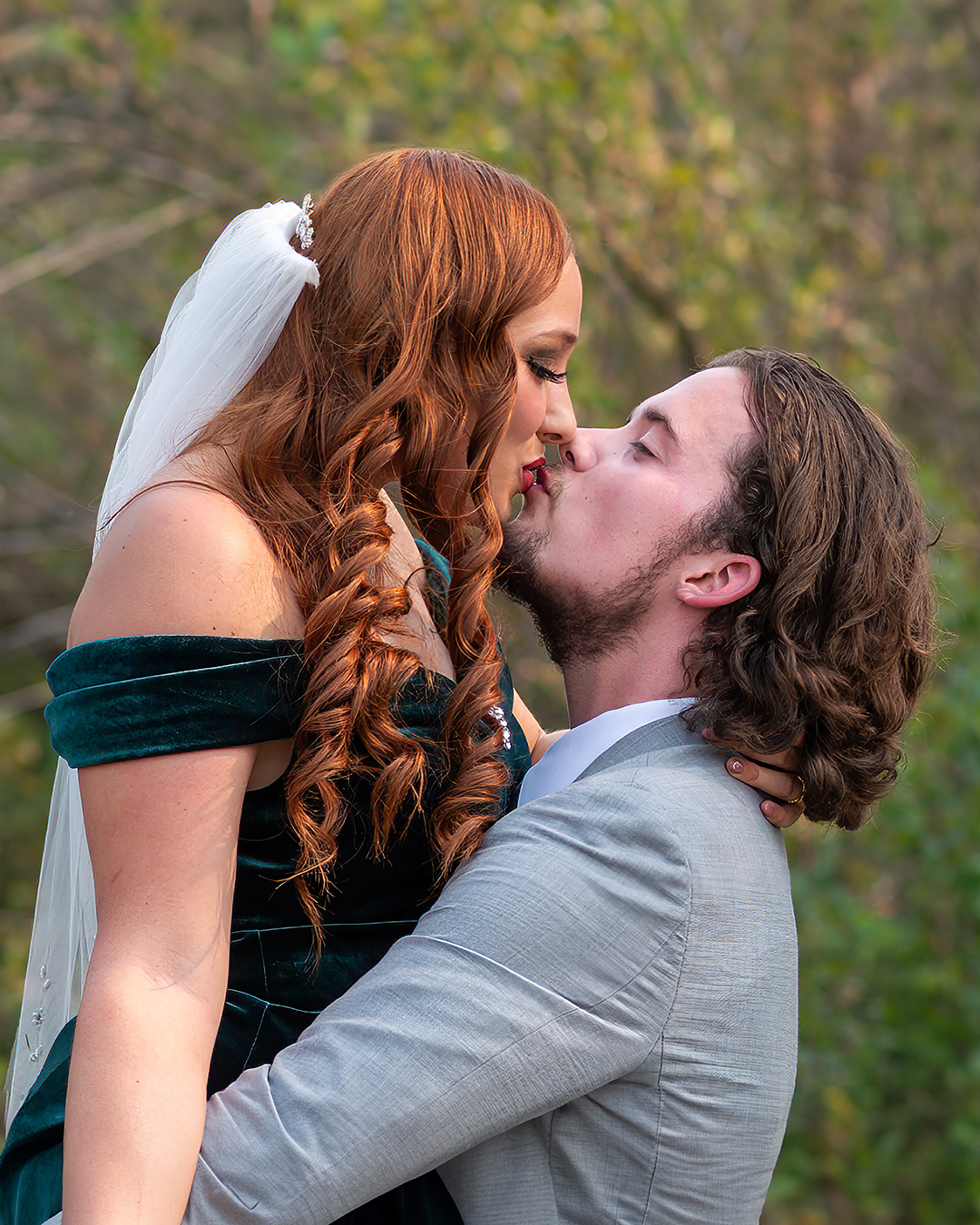 Bride kissing her groom on her wedding day. She is wearing a emerald green wedding dress. Groom is lifting the bride and wearing a gray suit. 