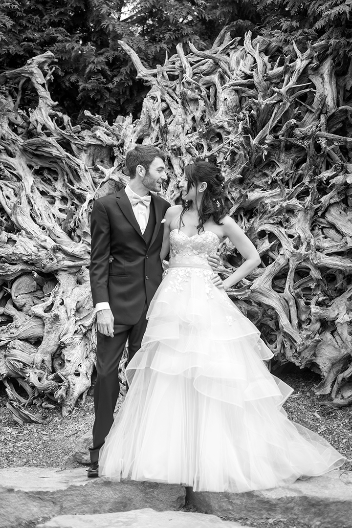 Couple stands in front of a fallen tree at the roots in the PNW. The photo is in black and white. The bride has a floral bodice with a tiered dress. While the groom is in a black tux. 