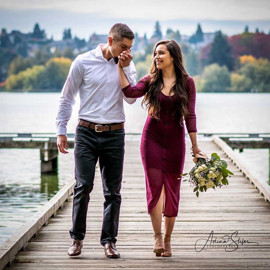 Couple walk hand in hand on a wooden dock in the PNW. The man kisses the woman's hand as she smiles while looking at the man. Woman is wearing a burgundy dress with tan sandals and carrying a white rose bouquet. The man is wearing black jeans with dress shoes and a white dress shirt.  