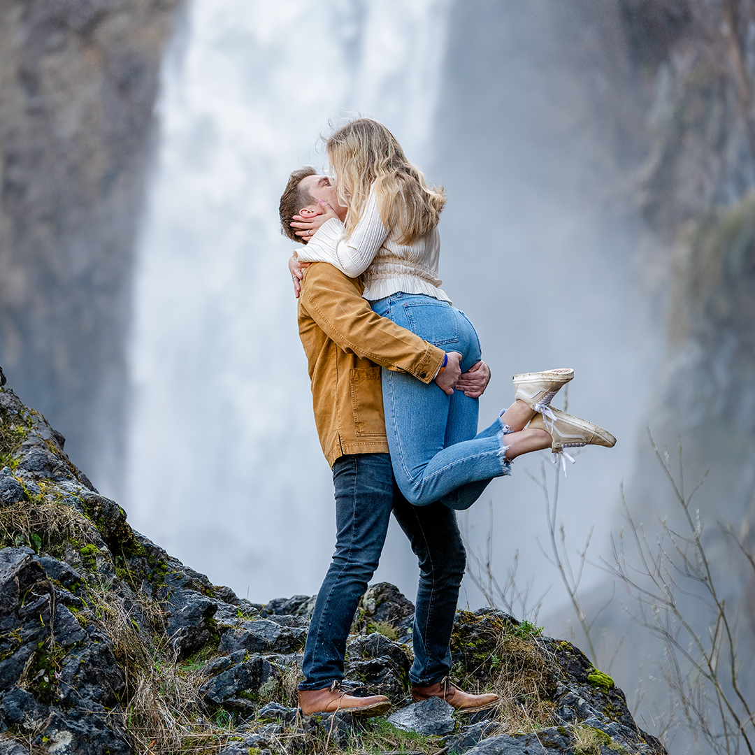 Couple kissing with a waterfall in the background while standing on a moss covered rock. 