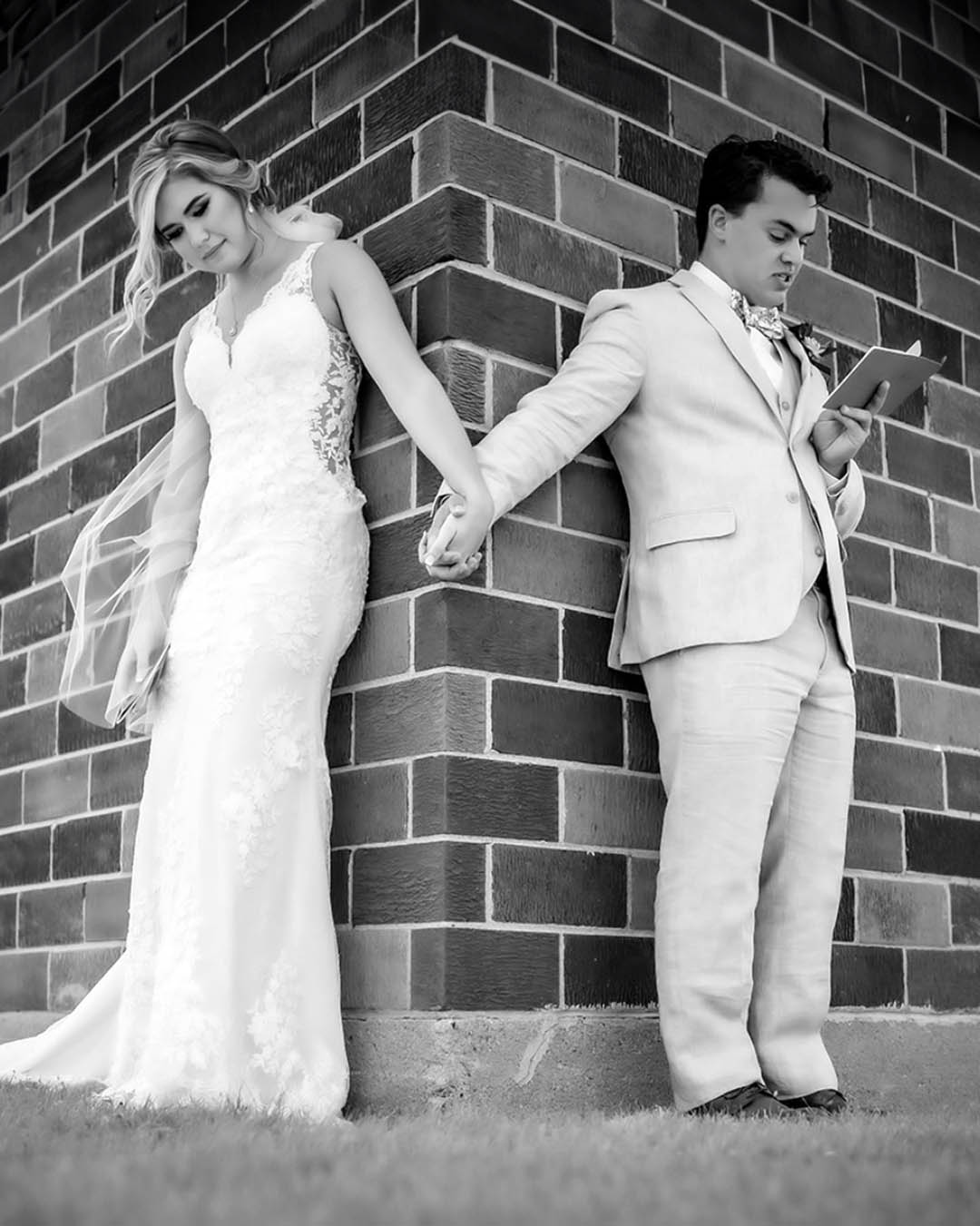 Black and white photo of a couple standing on either side of a corner of a brick wall while reading their personalized vows during a private first touch and first look moment during their wedding day. The bride and groom are holding hands and their vow books. The bride has a floor length veil. 