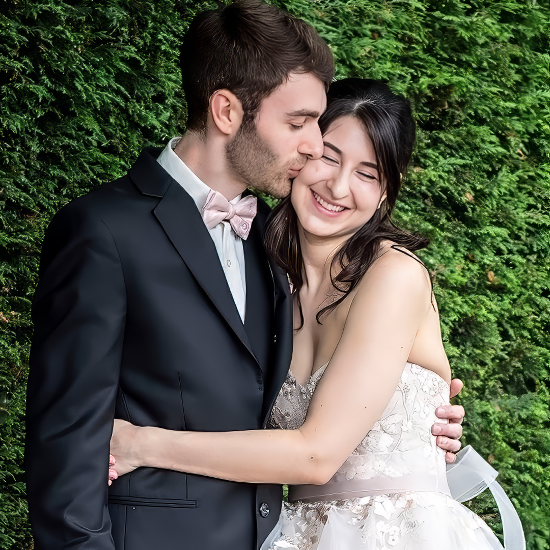 Quiet moment between groom and bride on their wedding day. Groom kisses bride's cheek in front of a green hedge behind them. 