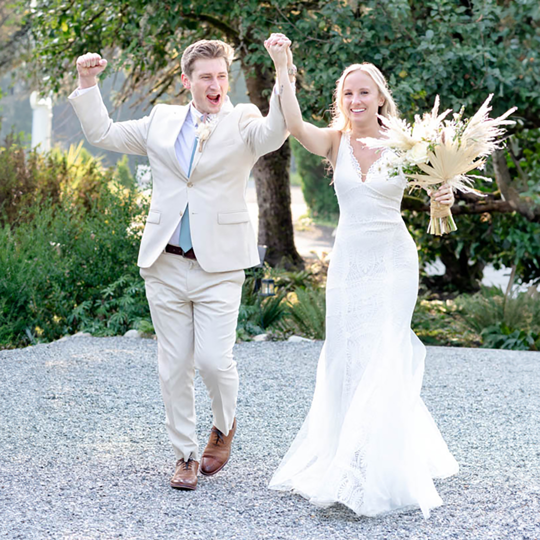 Bride and groom cheer as they enter the reception during their wedding day. Bride is holding a bouquet of flowers and groom is wearing a tan suit. 