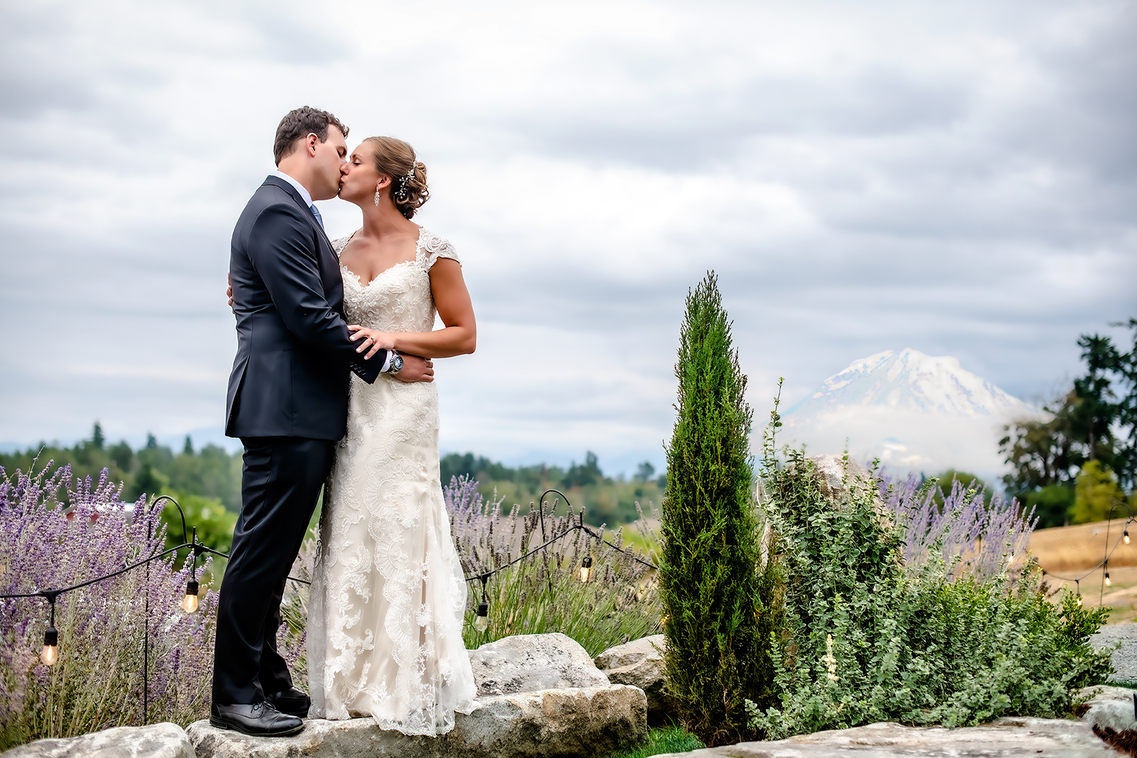 Bride and Groom stand on a rock overlooking a valley with the Cascade mountain range in the background. Bride and groom are kissing. Bride is wearing a lace covered gown and the groom is in a black tux near Portland Oregon.