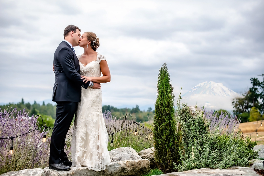 Bride and Groom stand on a rock overlooking a valley with the Mount Rainier in the background. Bride and groom are kissing. Bride is wearing a lace covered gown and the groom is in a black tux near Seattle, Washington.