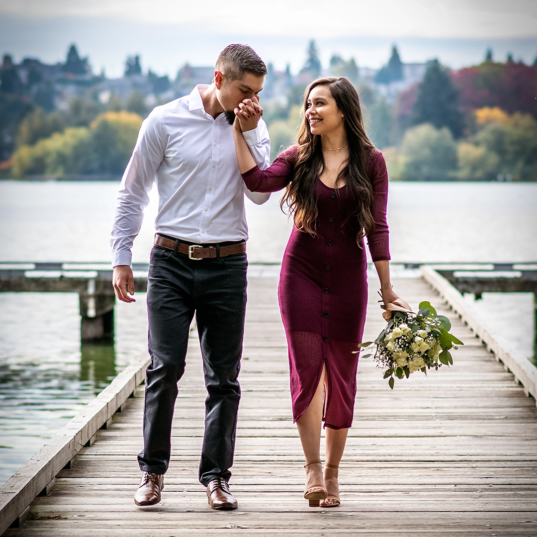 Couple walk hand in hand on a wooden dock in Seattle. The man kisses the woman's hand as she smiles while looking at the man. Woman is wearing a burgundy dress with tan sandals and carrying a white rose bouquet. The man is wearing black jeans with dress shoes and a white dress shirt.  