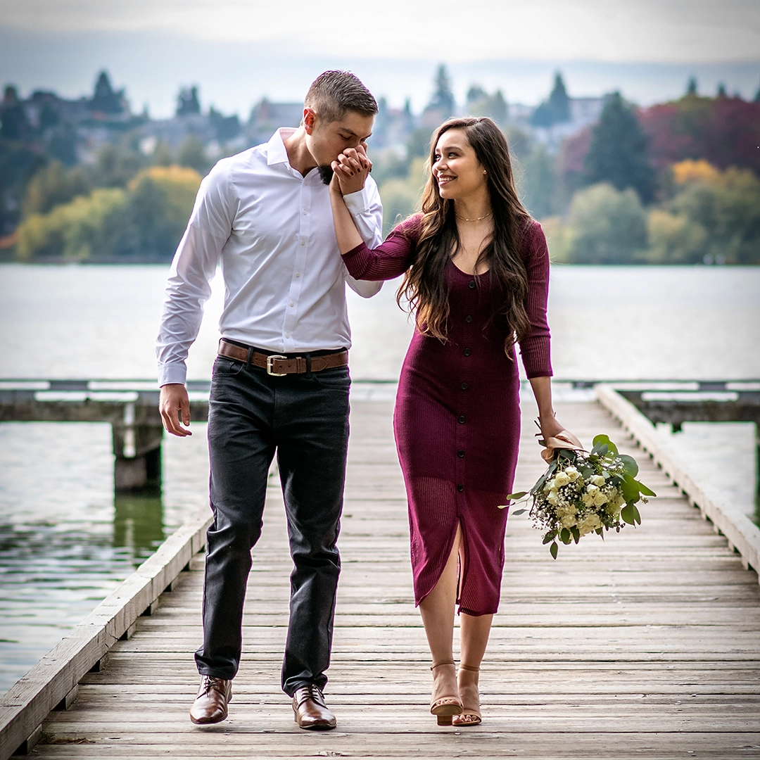 Couple walk hand in hand on a wooden dock in Maple Valley. The man kisses the woman's hand as she smiles while looking at the man. Woman is wearing a burgundy dress with tan sandals and carrying a white rose bouquet. The man is wearing black jeans with dress shoes and a white dress shirt.  