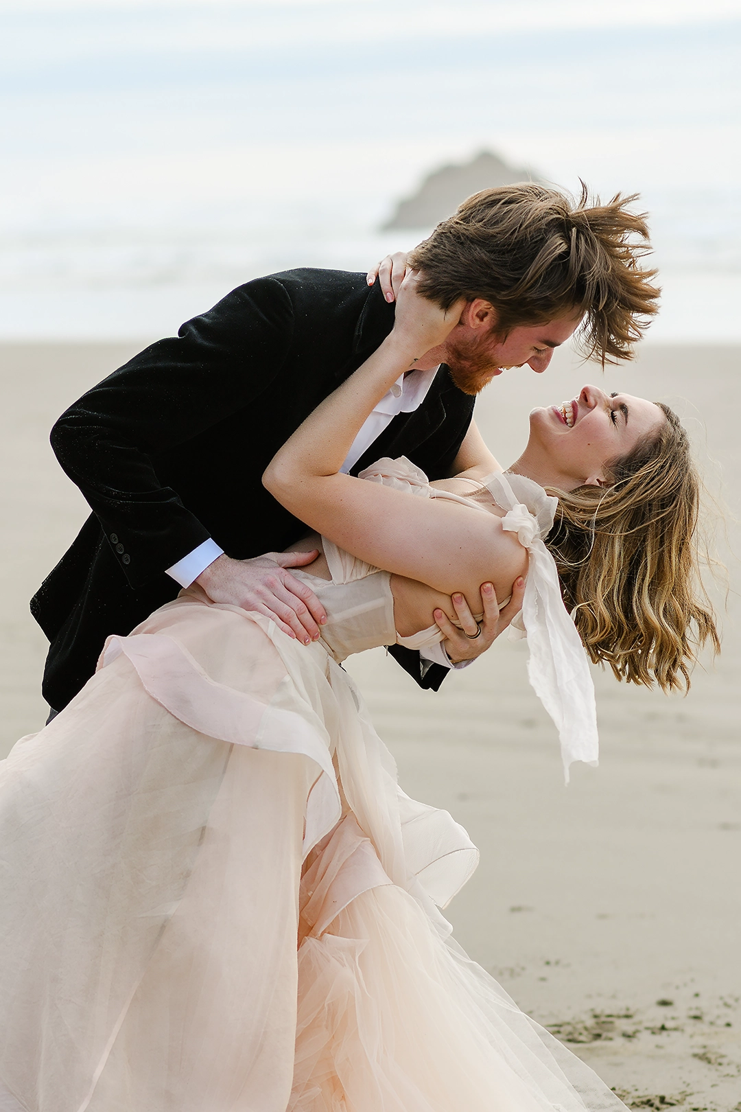 Couple laughs while the groom dips the bride back. The wind is blowing their hair on the beach. Rocks jut out of the ocean in the background.