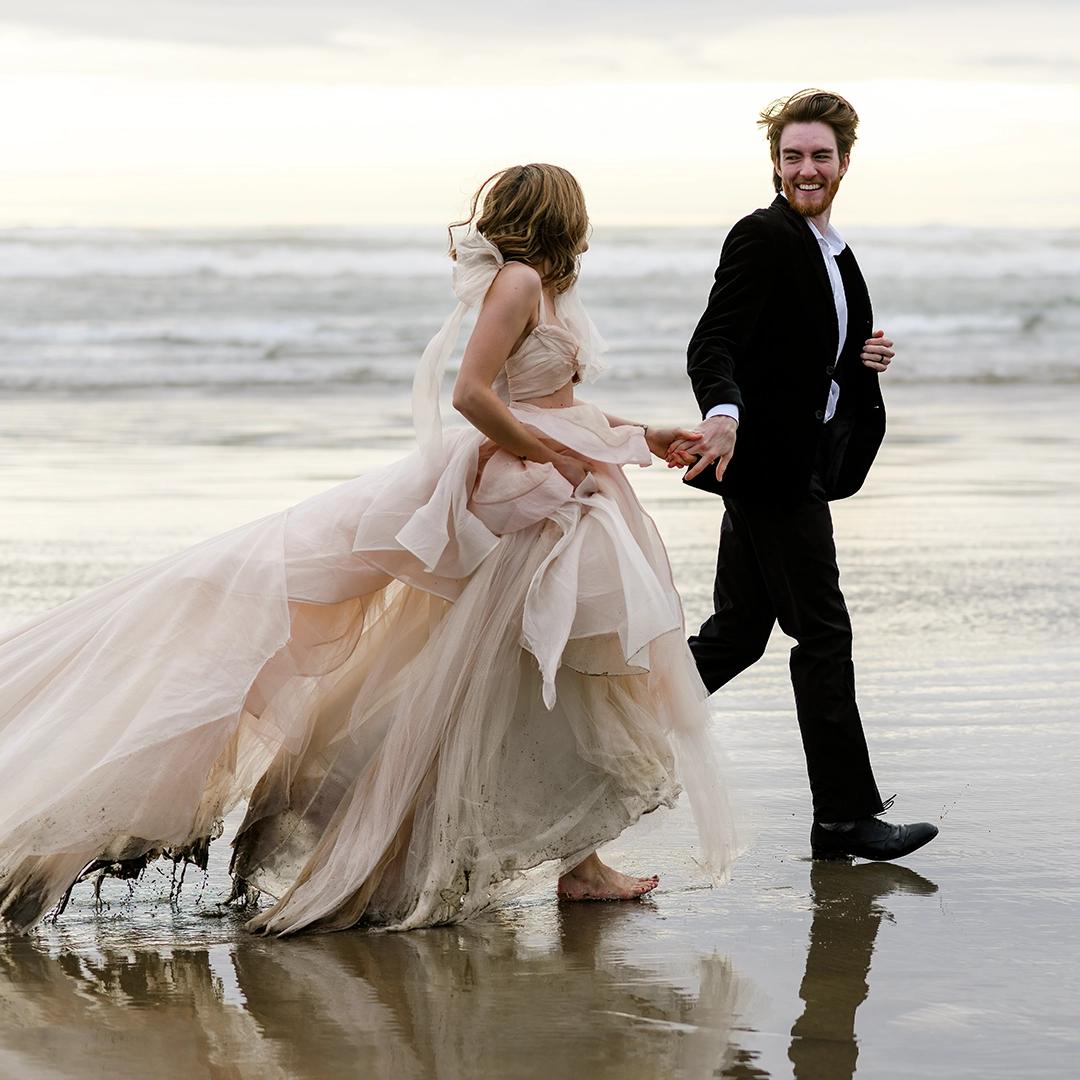 A couple runs on the beach at Cannon Beach, Oregon. They are hand in hand. Water drips from the brides dress. She has it gathers in her arms. The couple is smiling while they run. 