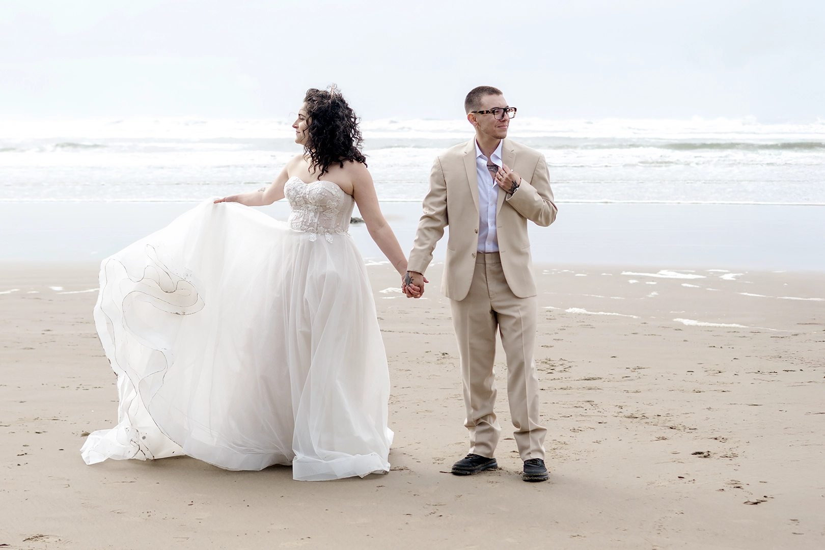 Couple stand hand in hand with the Pacific Ocean behind them on Cannon Beach. Bride is swinging her dress while groom is in a tan suit looks the opposite direction. 