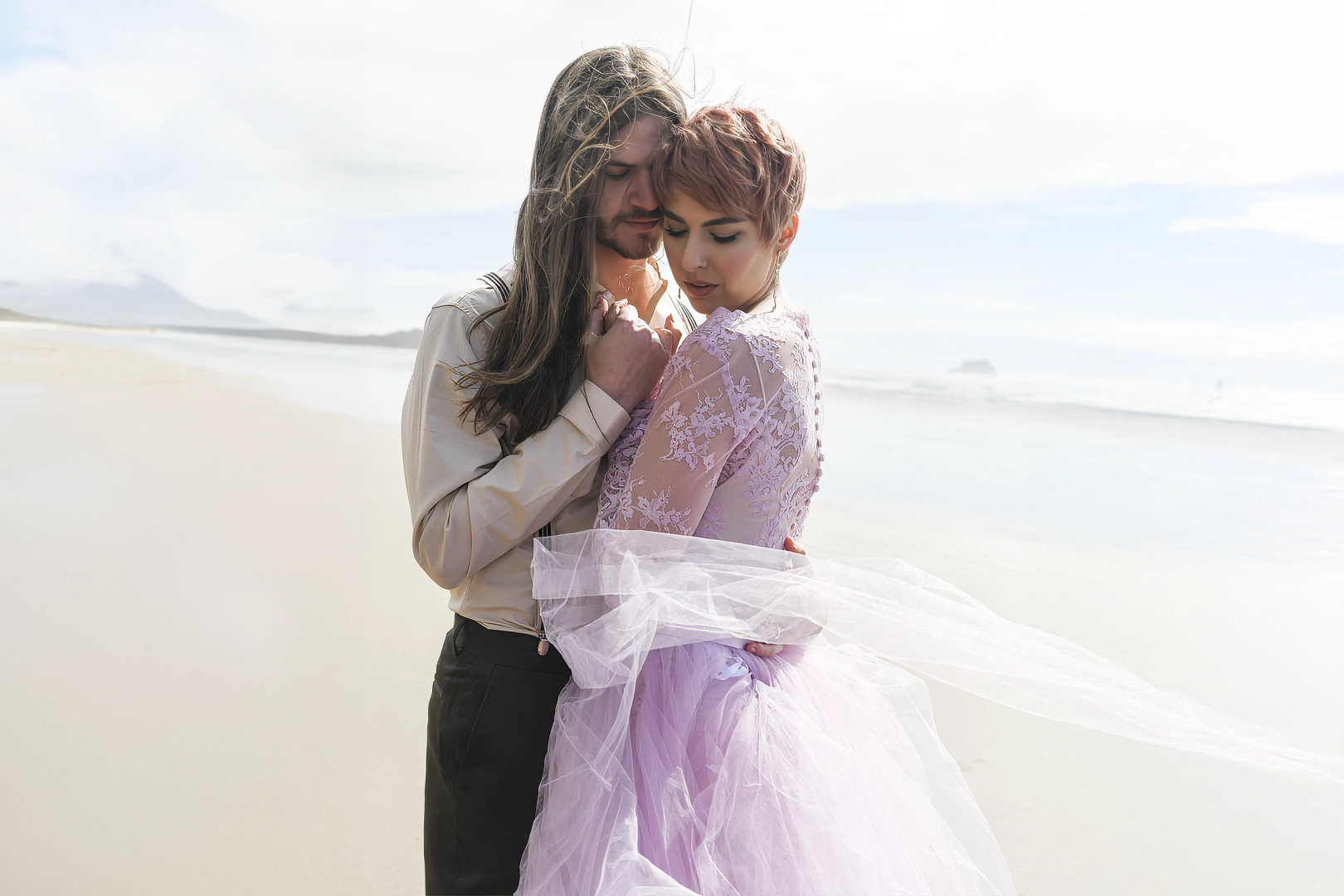 Bride and groom along the shore of Cannon Beach. Both are looking down with the wind blowing their hair and clothes. 