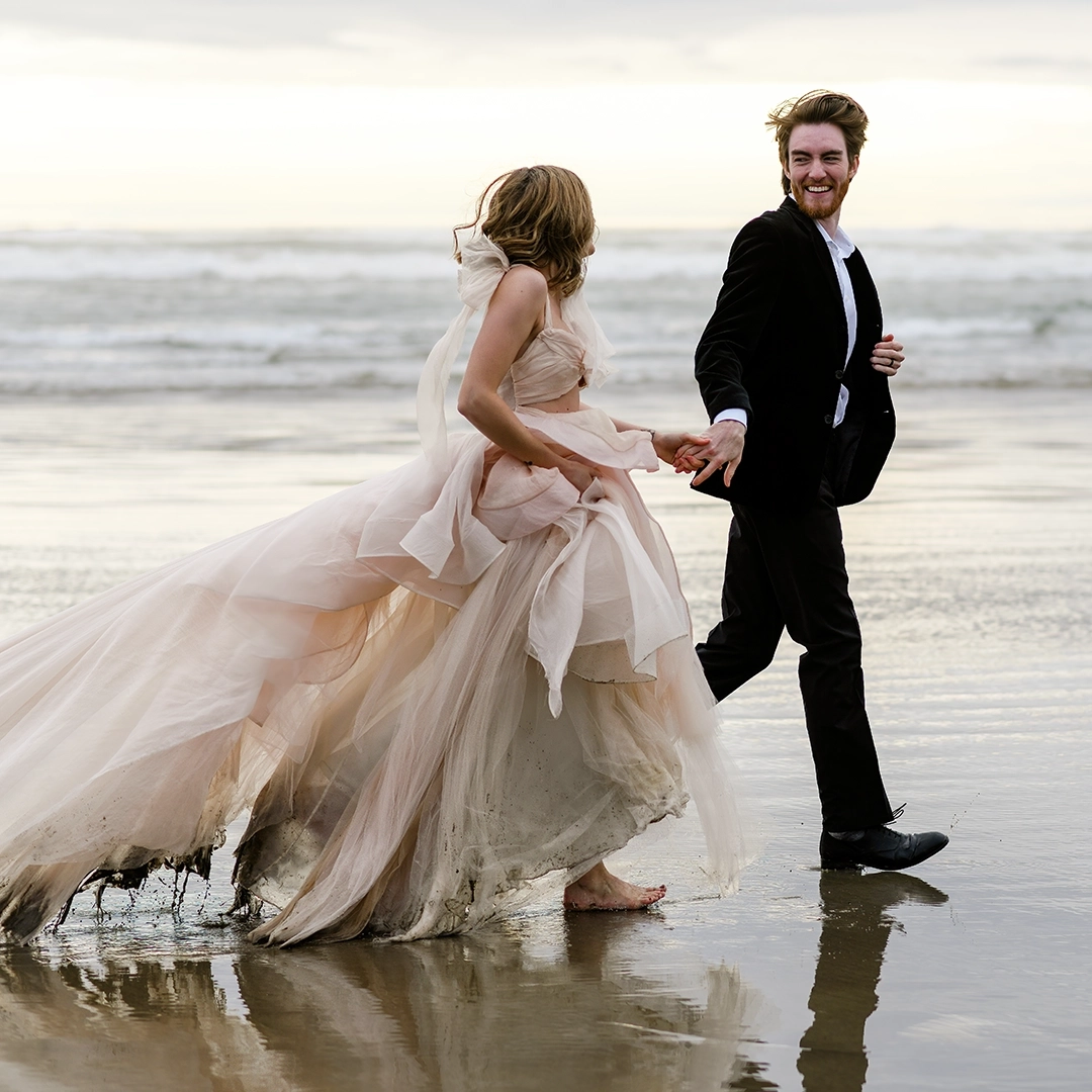 A couple runs on the beach at Cannon Beach, Oregon. They are hand in hand. Water drips from the brides dress. She has it gathers in her arms. The couple is smiling while they run. 