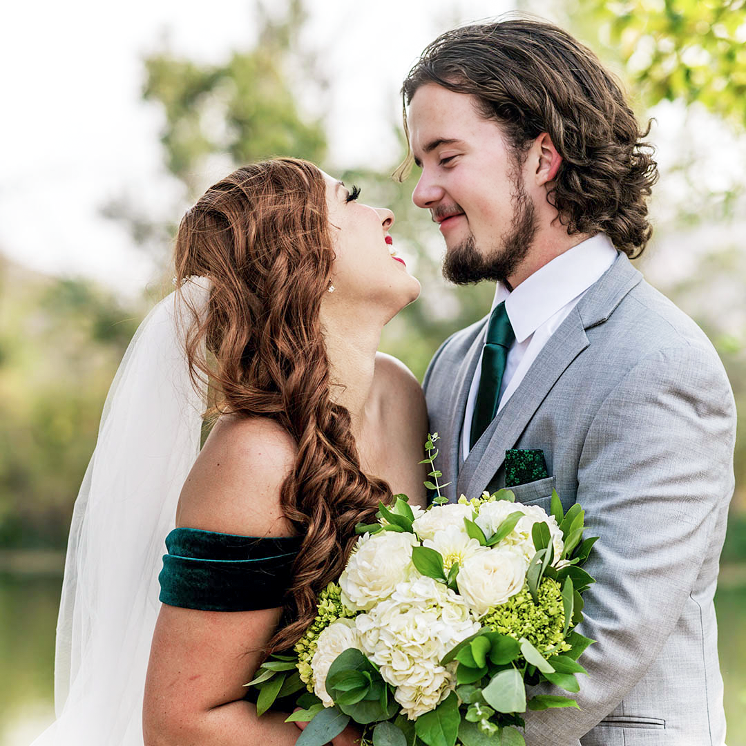 A couple stands close together with big smiles on their faces. The bride is wearing an emerald green wedding dress with a cathedral length veil and holding a floral bouquet of white chrysanthemums and green leaves and florals. The groom is wearing a grey suit with an emerald green tie and pocket square.