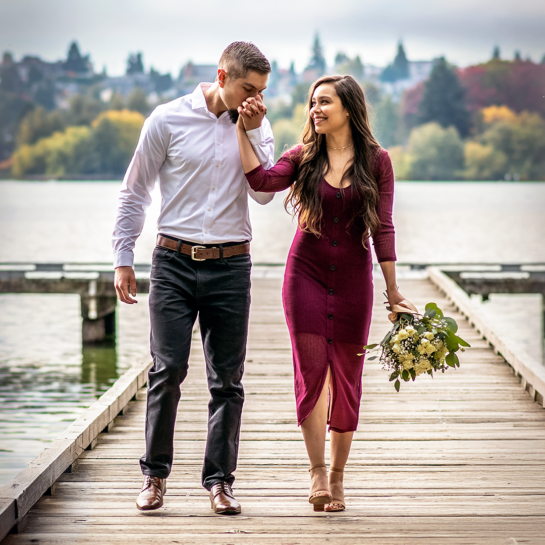 Couple walk hand in hand on a wooden dock in Seattle. The man kisses the woman's hand as she smiles while looking at the man. Woman is wearing a burgundy dress with tan sandals and carrying a white rose bouquet. The man is wearing black jeans with dress shoes and a white dress shirt.  