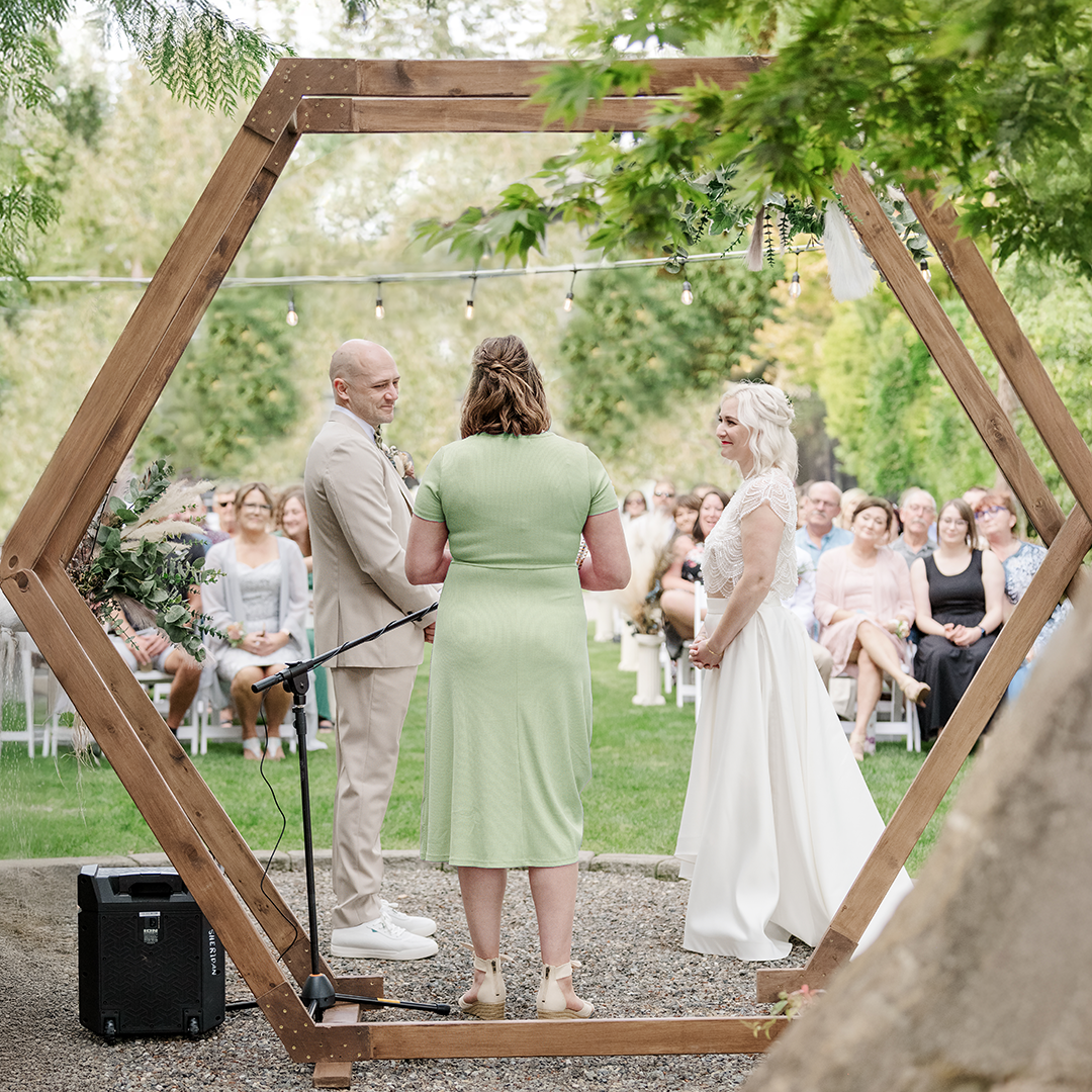 A graceful bride and groom exchange vows under a wooden arch adorned with greenery, surrounded by guests, her elegant white dress and his light suit reflecting the serene beauty of a wedding celebration.