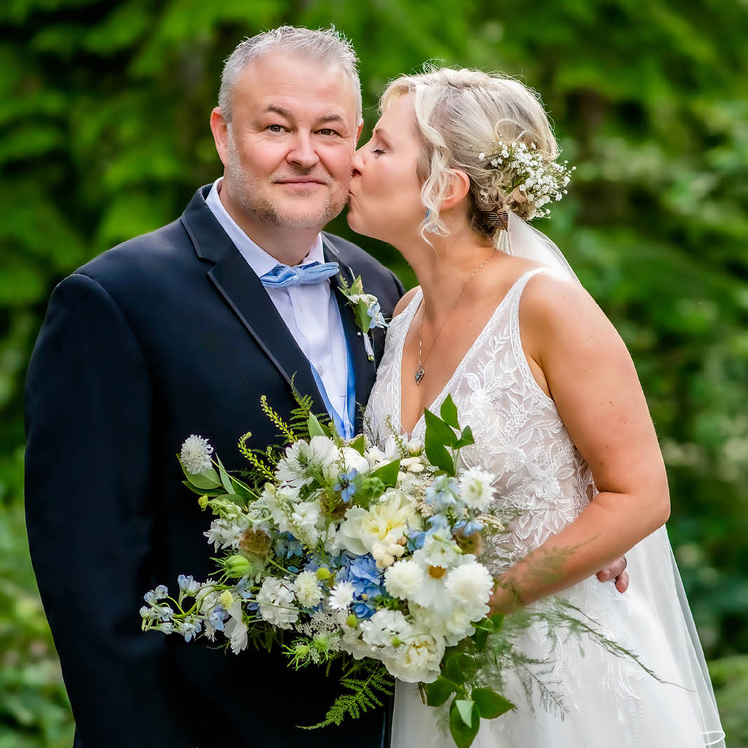 A sophisticated bride and groom share a loving kiss amidst lush greenery, she in an embroidered gown with a bouquet, he in a sleek suit, highlighting the intimate allure of a wedding portrait.