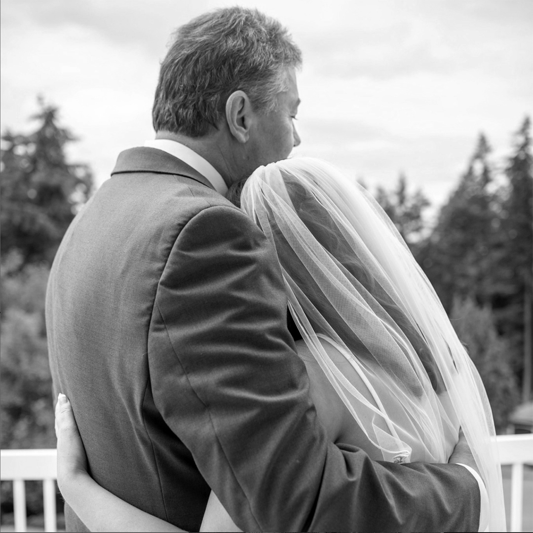A heartfelt embrace between a bride in a flowing veil and her dad in a tailored suit, set against a scenic backdrop of trees, capturing the emotional depth of her wedding..