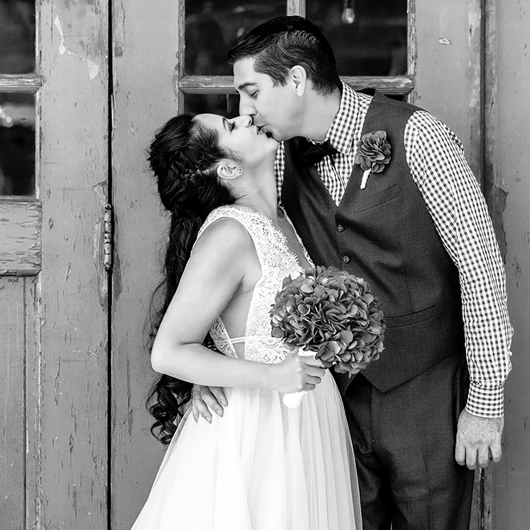 A stylish couple shares a passionate kiss against a rustic wooden door, the bride in a lace gown with a bouquet of hydrangeas, embodying the romantic charm of a wedding ceremony.