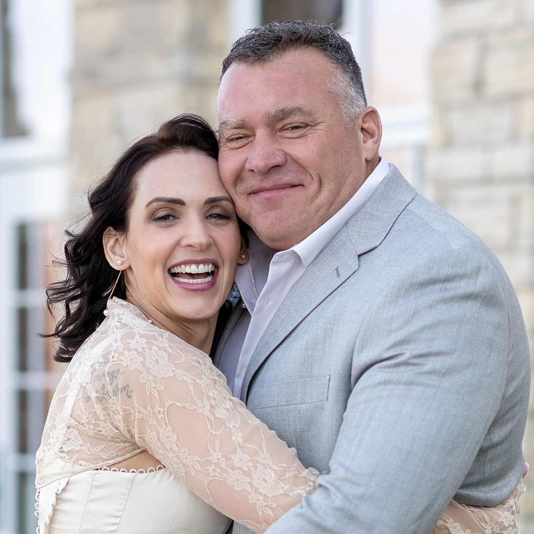 A joyful couple embraces closely, with the woman in a lace wedding dress and the man in a light gray suit, standing in front of a stone building.