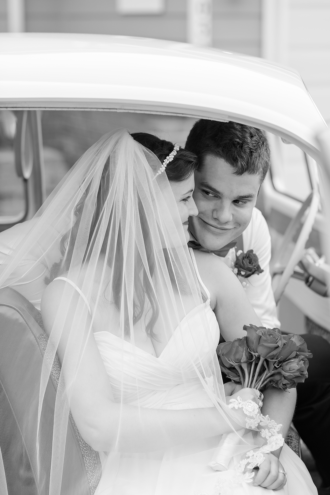 Black and white photo of a bride and groom sitting closely together in a vintage car. The bride, wearing a white wedding dress and veil, holds a bouquet of roses, while the groom, in a suit with a boutonniere, looks at her affectionately.