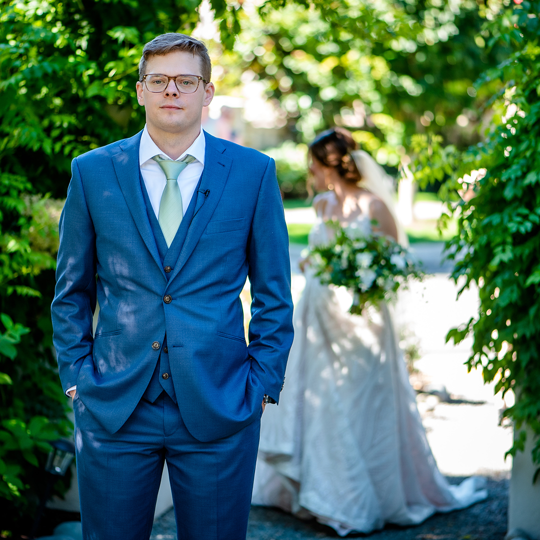 A graceful bride and groom exchange vows under a wooden arch adorned with greenery, surrounded by guests, her elegant white dress and his light suit reflecting the serene beauty of a wedding celebration.