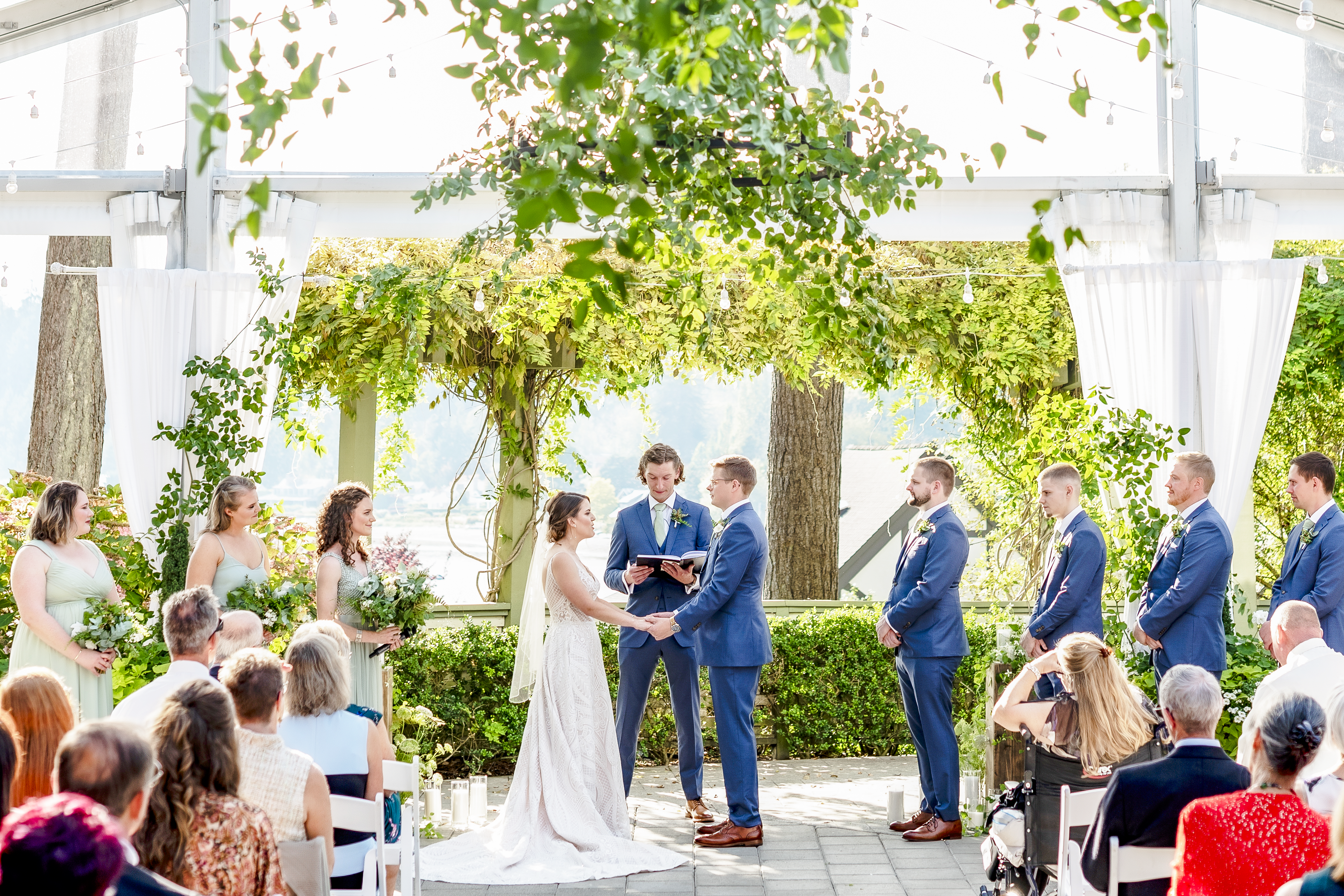 Garden wedding ceremony where couple holds hands while the officiant speaks and guests look on with the wedding party on each side of the aisle.