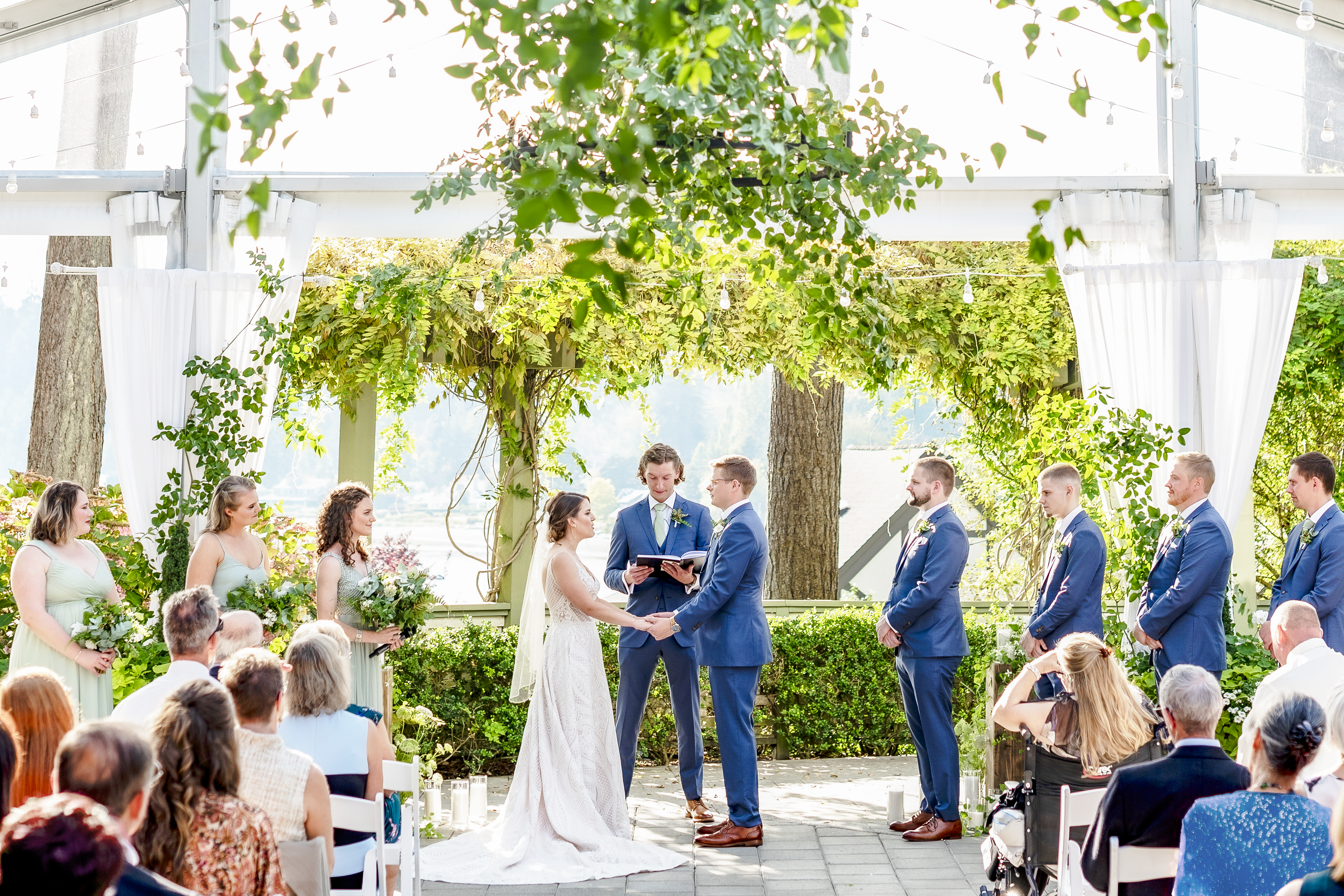 Garden wedding ceremony where couple holds hands while the officiant speaks and guests look on with the wedding party on each side of the aisle.