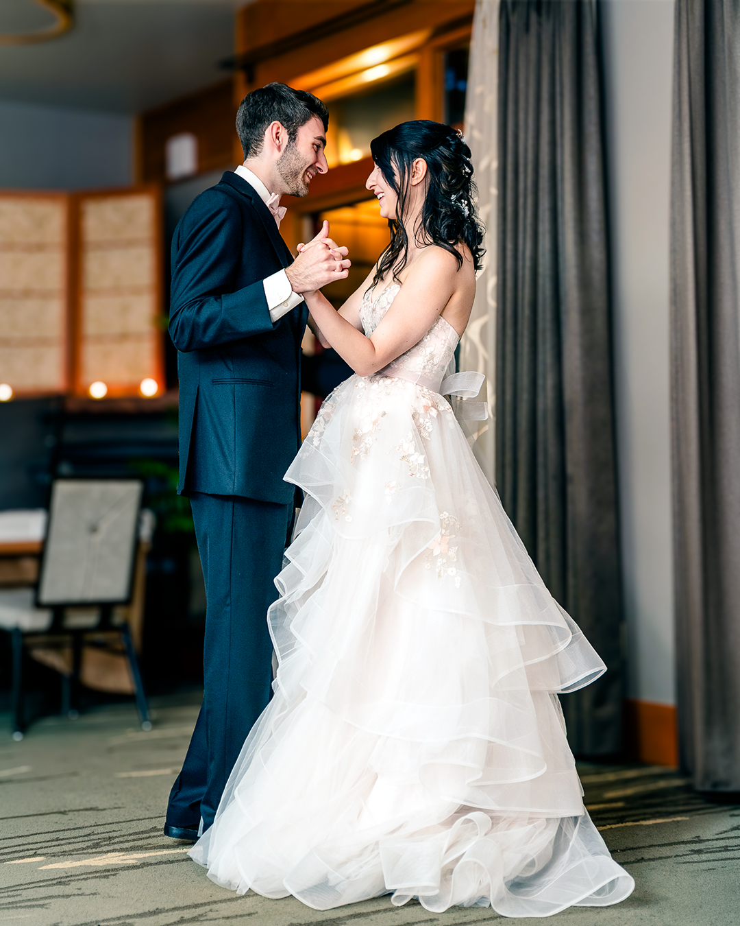 Couple holds hands while dancing their first dance on their wedding day in a dark banquet hall.