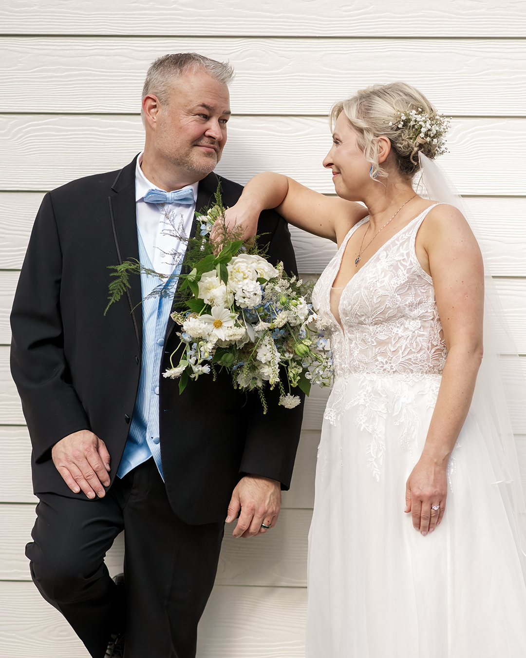 Bright, harsh light wedding day. Couple stands against a white barn wall while looking at each other with love.