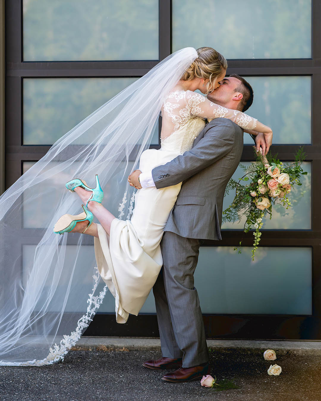 A couple kisses while the groom lifts the bride up in front of frosted garage doors on their wedding day. The bride is wearing cute blue shoes.
