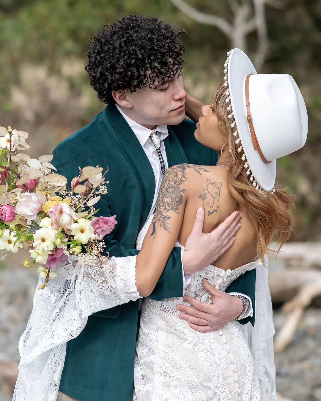 A modern-classic couple shares an intimate moment, the bride in a lace wedding dress with a tassled white hat, and the groom in a tailored green suit jacket, embraces on Cannon Beach, OR during their intimate wedding celebration.