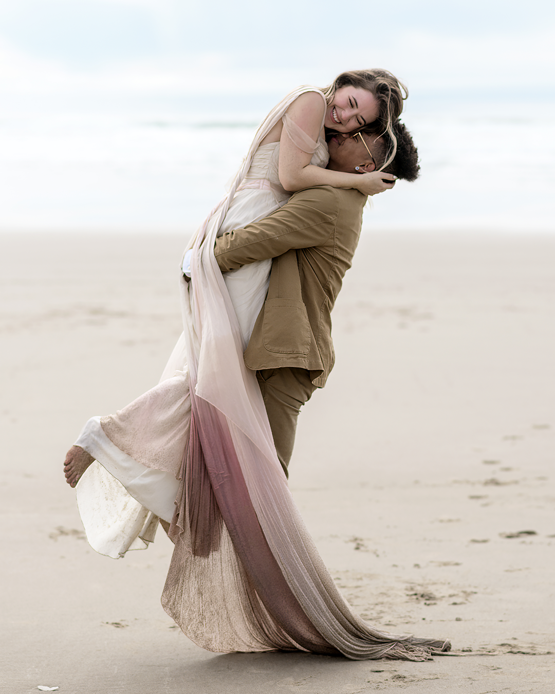 A groom lifts his bride while kissing her on the cheek while lifting her up as he stands on the Oregon Coast. She is wearing a cream and pink wedding dress and he is in a tan colored suit. They are both barefoot.