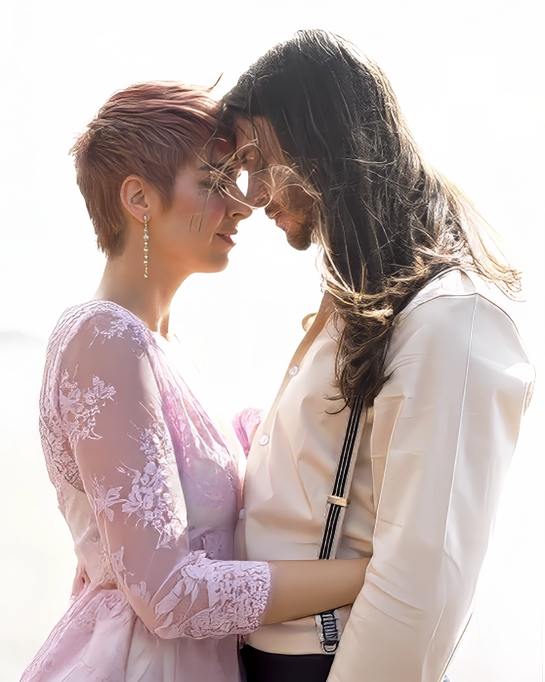 A couple stands touching foreheads on the beach on their wedding day. Bride is wearing a pink wedding dress while the groom is wearing a brown and tan suit with suspenders. Their hair blows in the breeze.