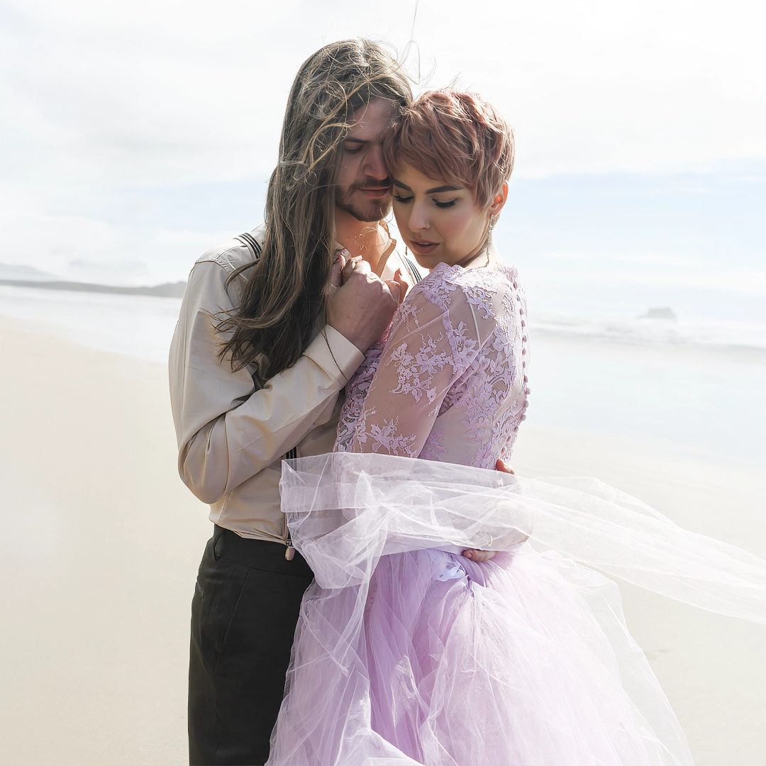 Bride and groom along the shore of Cannon Beach. Both are looking down with the wind blowing their hair and clothes. 
