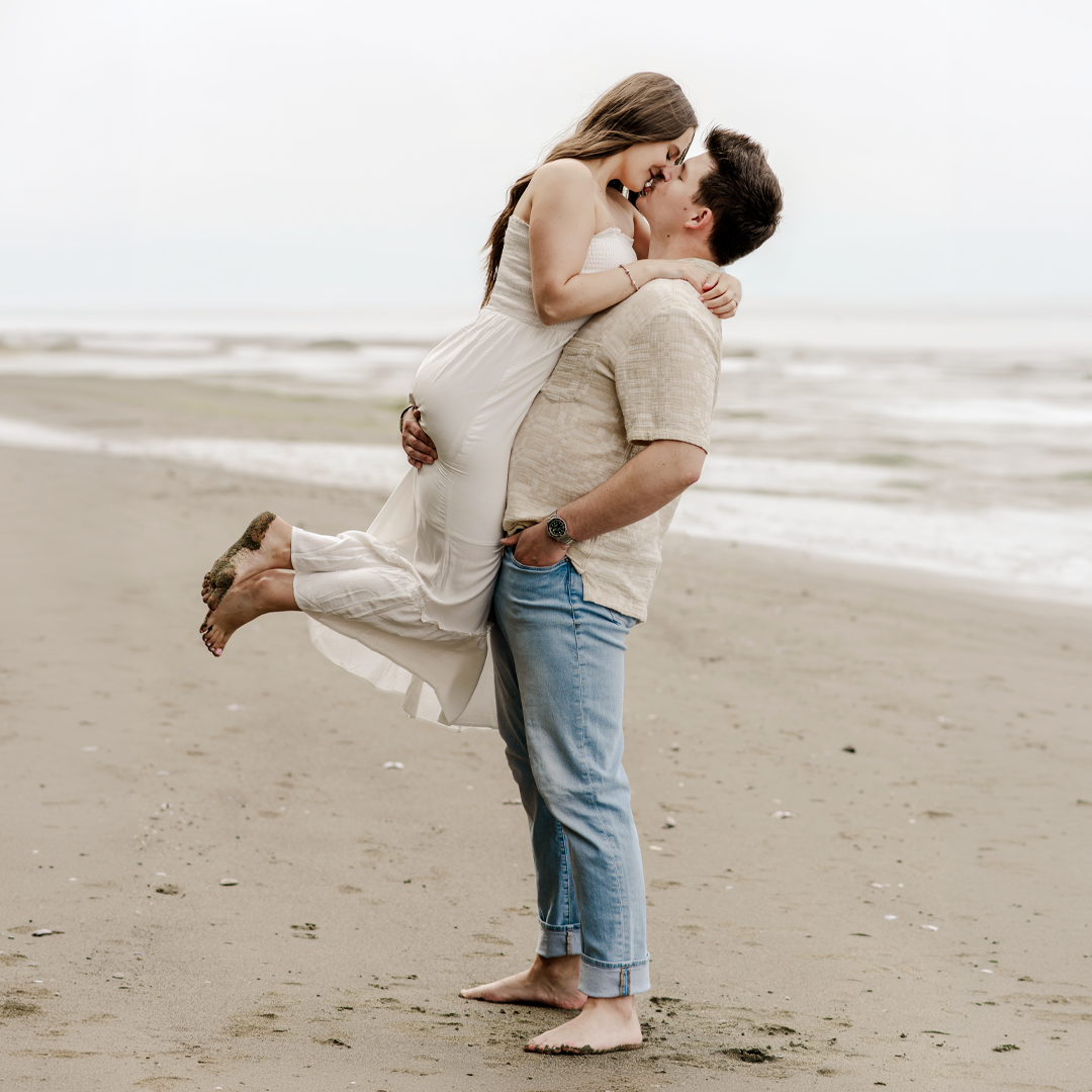 Groom lifts his future bride in a lifted embrace. Her sandy feet raise up at the knees. Both bride and groom are barefoot. Man is wearing jeans, woman is wearing a cream colored dress. 