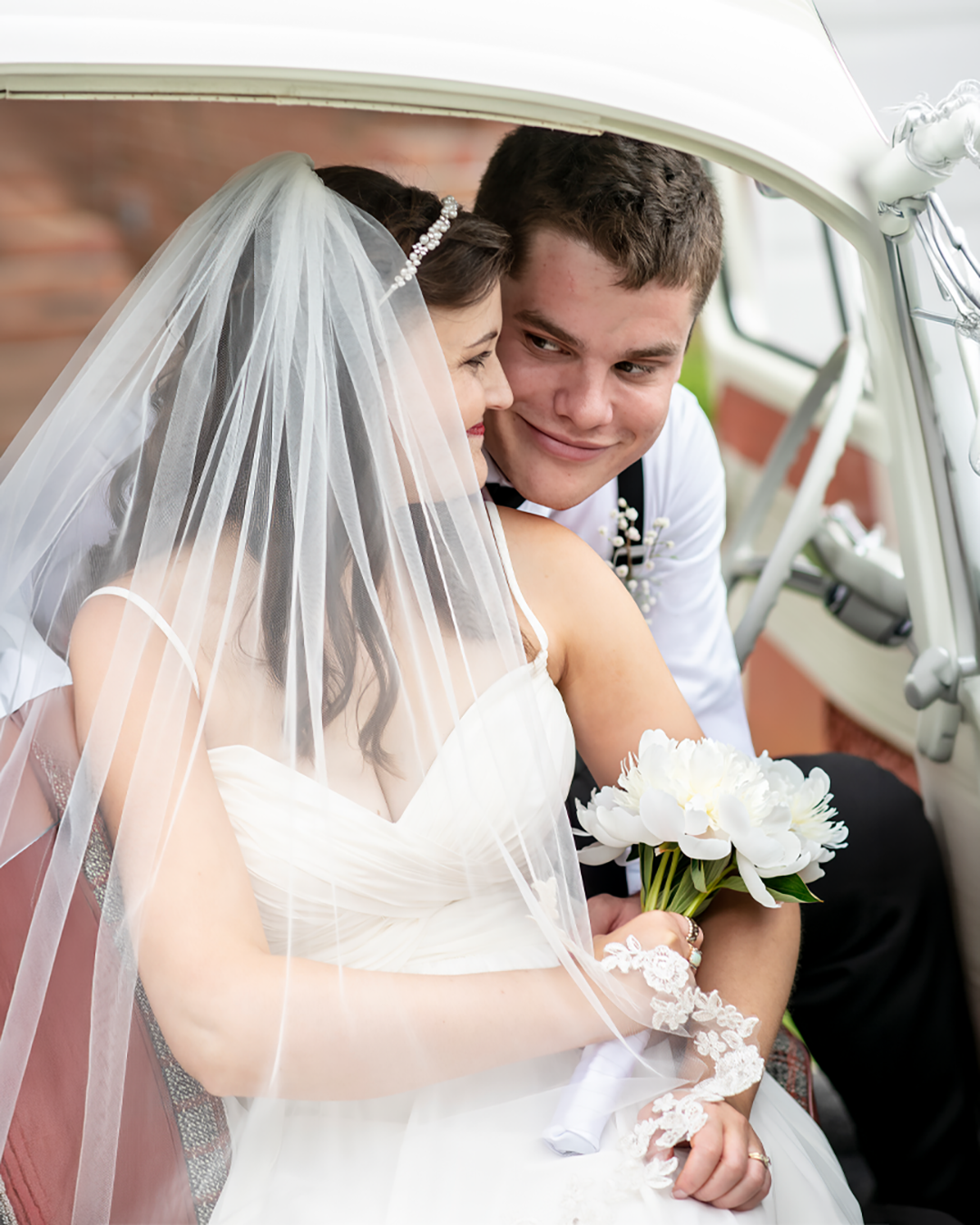 Couple sits in a VW Bug car at their wedding in Maple Valley. They are looking at each other while the bride holds a bouquet of white peonies.