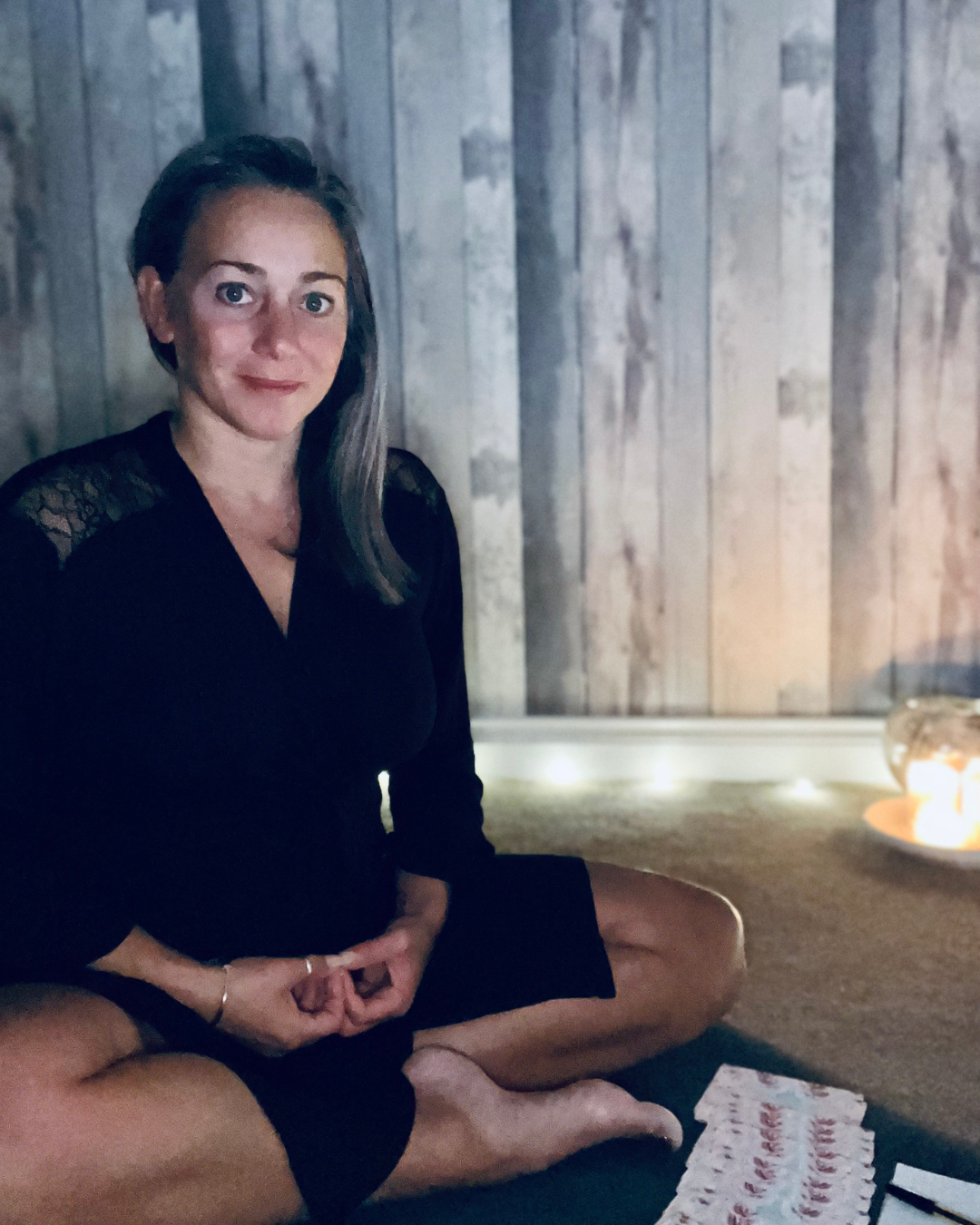 Lee-ann Cordingley sitting cross-legged on the floor in a candlelit room, wearing a black wrap top, hands resting in a meditative gesture. A soft warm light glows beside her, evoking the embodied "dark yoga" practice she describes - free movement, candles, and coming home to the body.