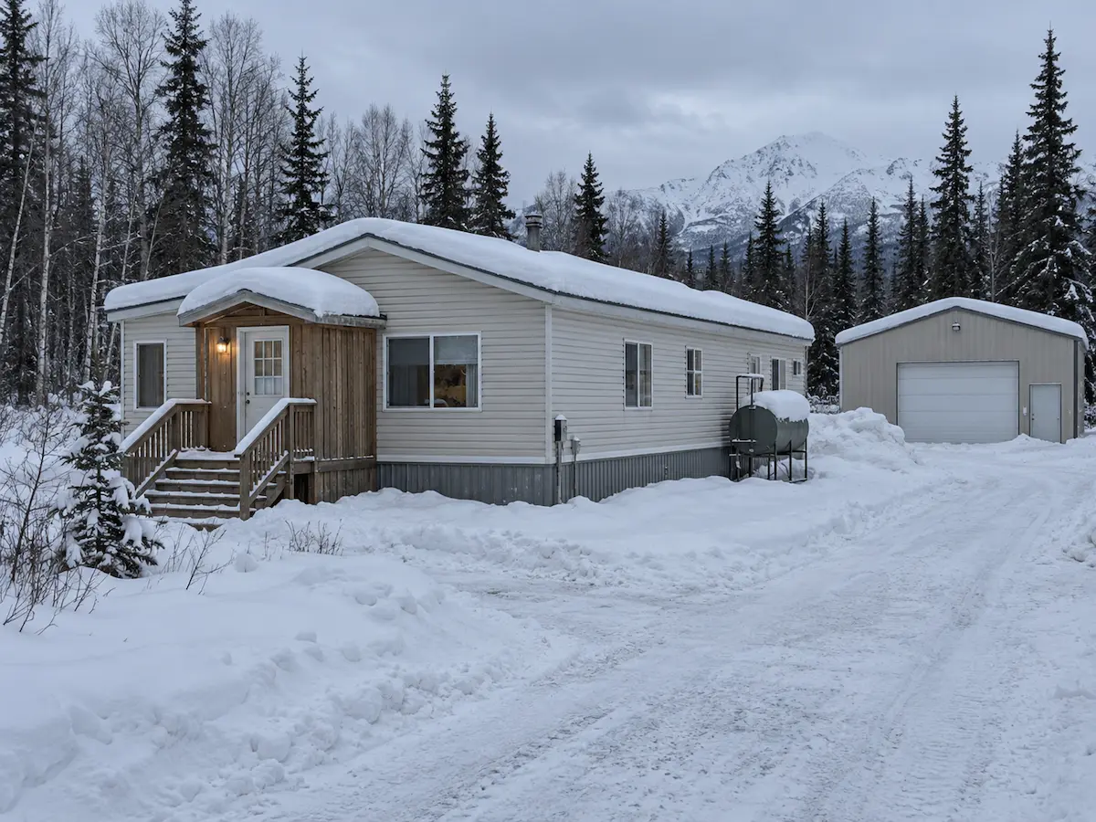 A single-wide manufactured home with an arctic entry, fuel tank and detached metal garage on a snow-covered rural lot in Southcentral Alaska
