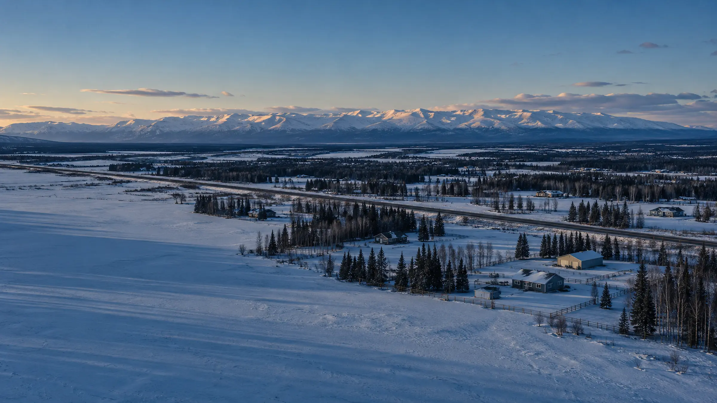 Rural homes and shops on large snow-covered lots along a highway in a wide Southcentral Alaska valley, with a snow-covered mountain range on the horizon