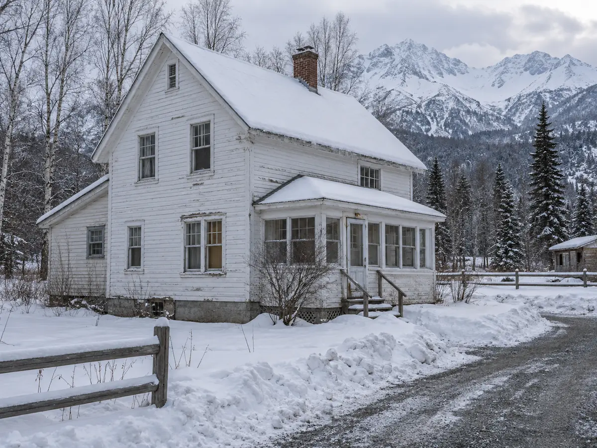 A weathered 1930s white clapboard farmhouse with an enclosed entry porch and brick chimney, in snow below the Chugach Mountains