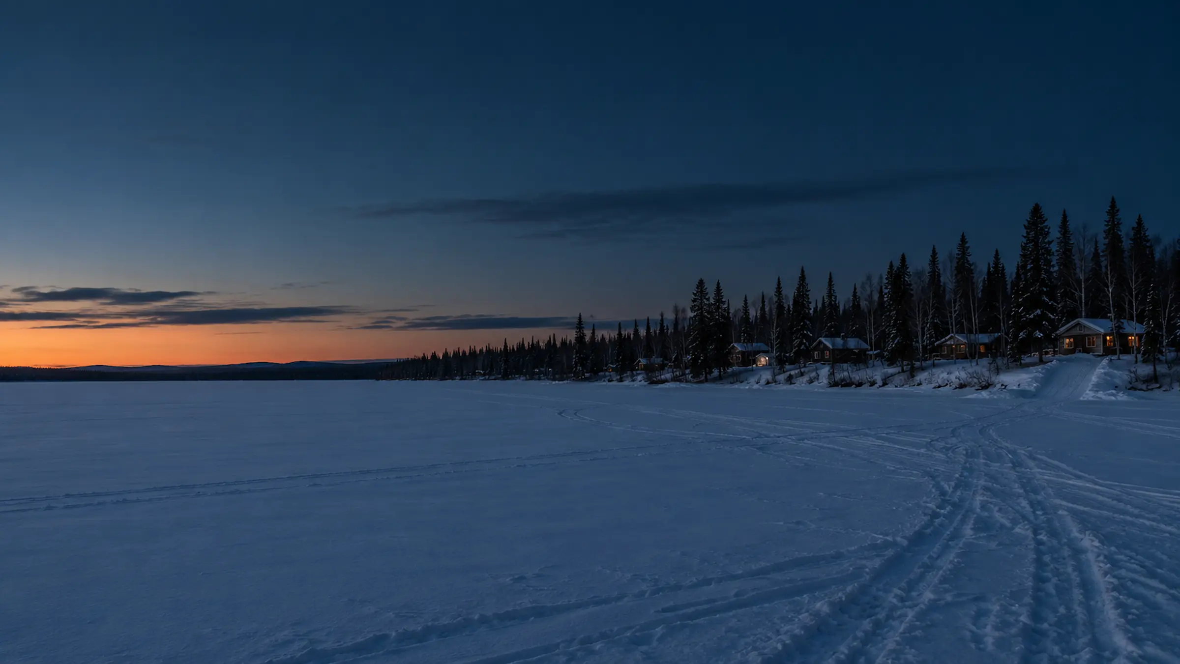 Cabins along the spruce shoreline of a frozen lake at dusk, with snowmachine tracks across the ice, Southcentral Alaska