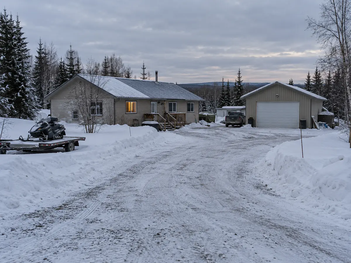 A single-story rural home with a detached garage, plowed driveway and a snowmachine on a trailer, on flat wooded ground in Southcentral Alaska