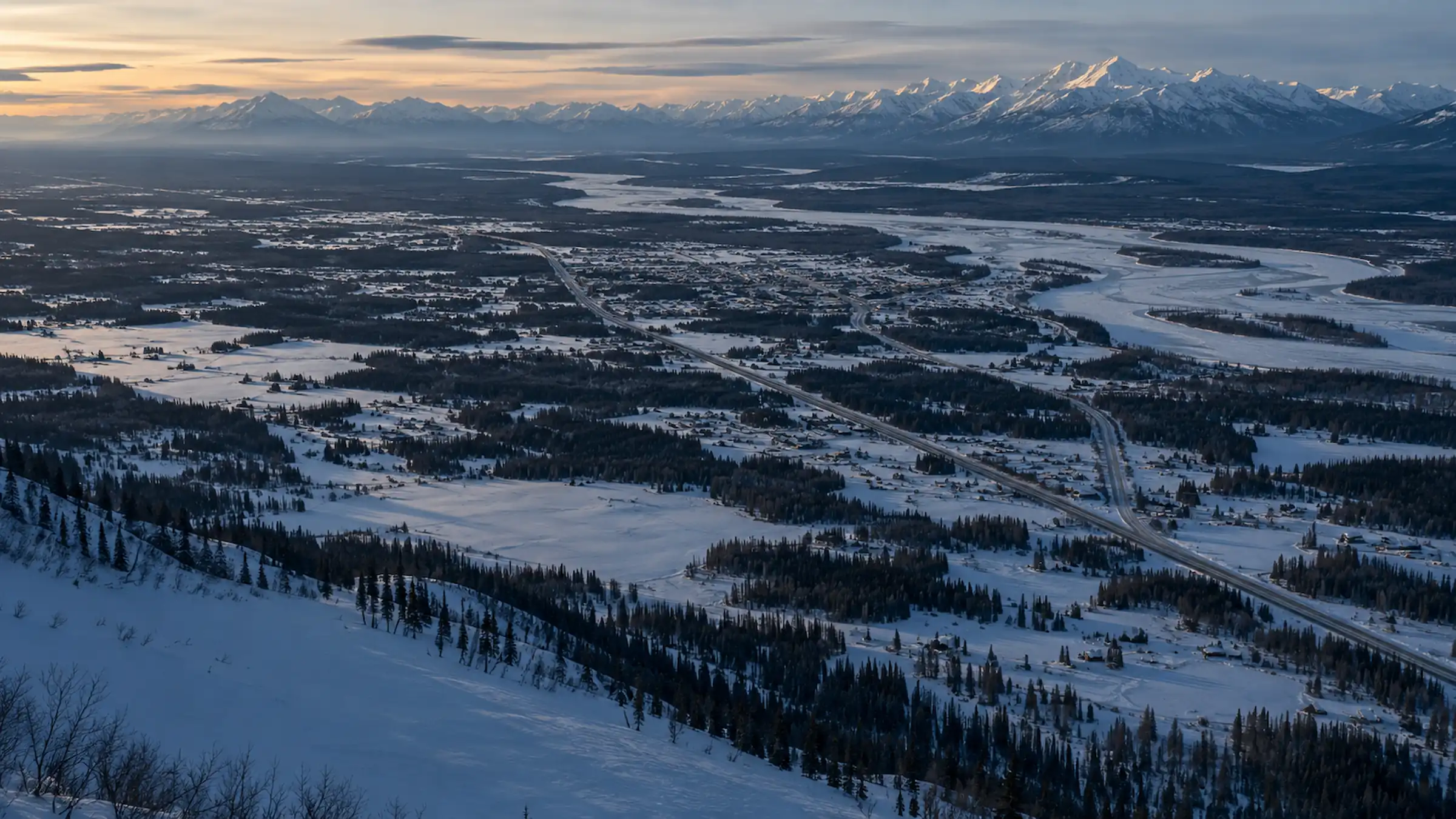 Aerial view across a broad snow-covered valley in Southcentral Alaska, with a highway, scattered homes, a braided frozen river and a long mountain range on the horizon