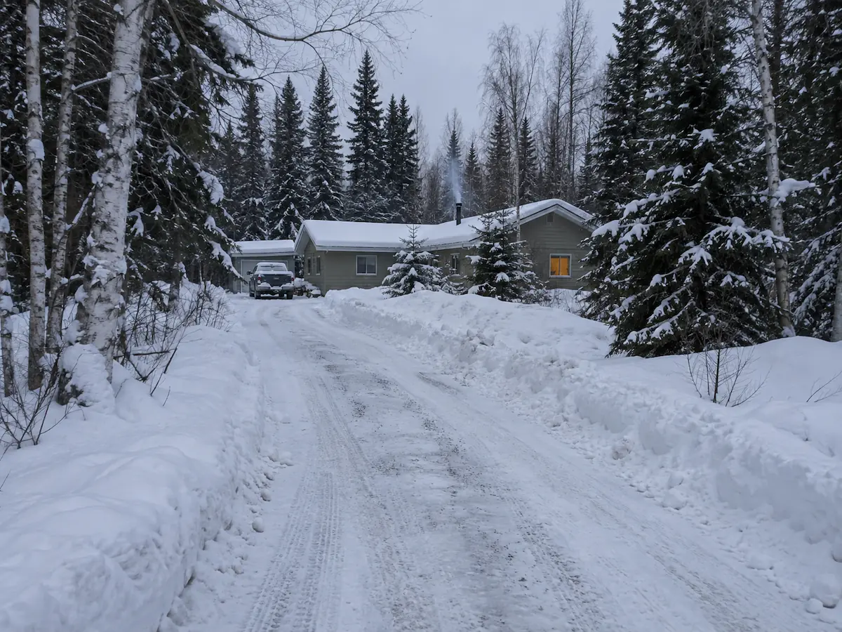 A plowed rural driveway between deep snow berms leading to a home with woodstove smoke, surrounded by spruce and birch in Southcentral Alaska